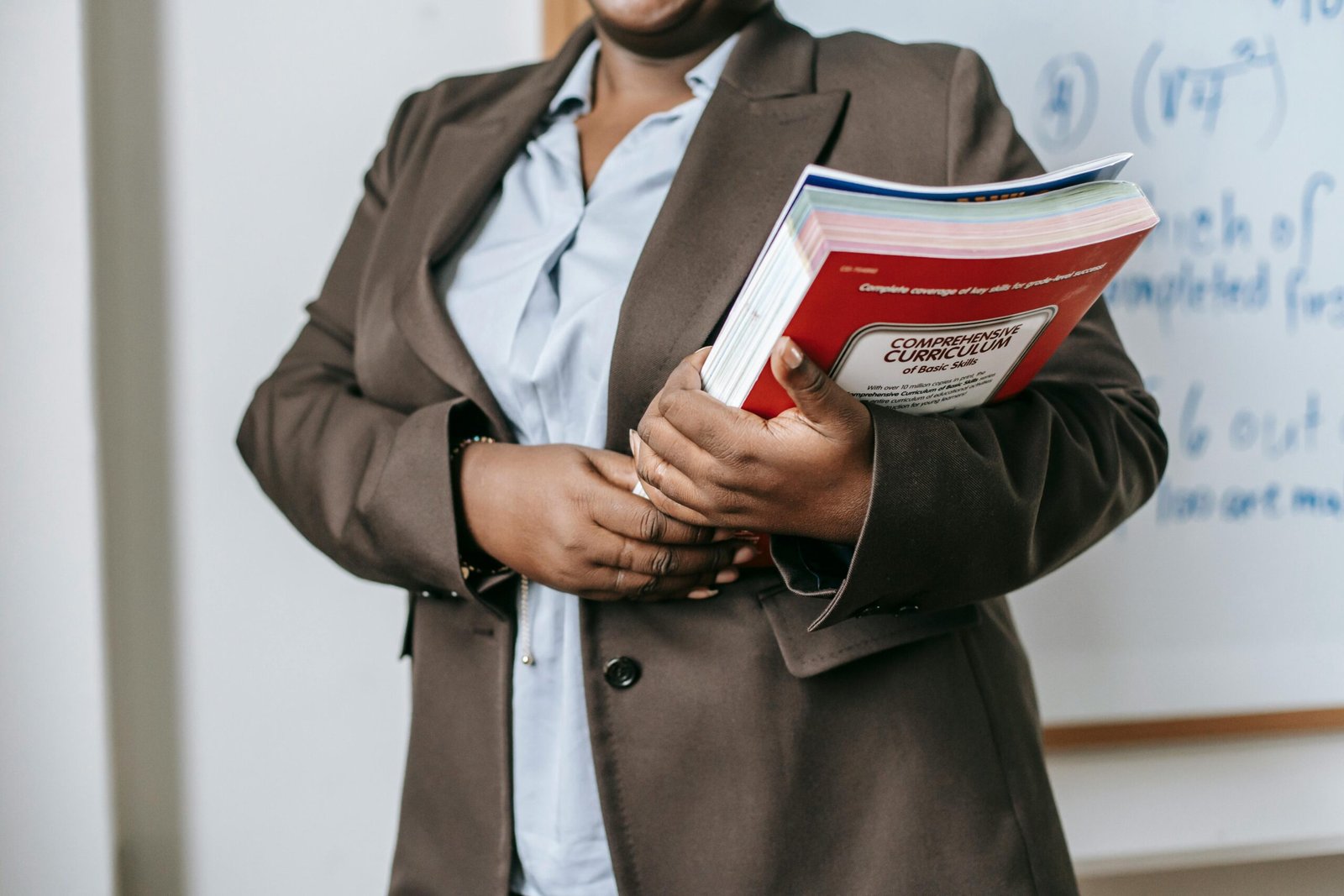 A professional educator in a formal setting holding a curriculum book in front of a whiteboard.